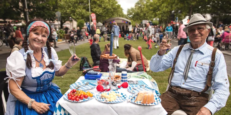 Ein älteres Pärchen sitzt in bayerischer Tracht an einem Picknick-Tisch auf Rollrasen und prostet dem Fotografen mit Sekt zu.
