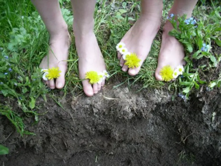 Blüten von Löwenzahn und Gänseblümchen schmücken Füße, die auf Gras und schwarzer Erde stehen.