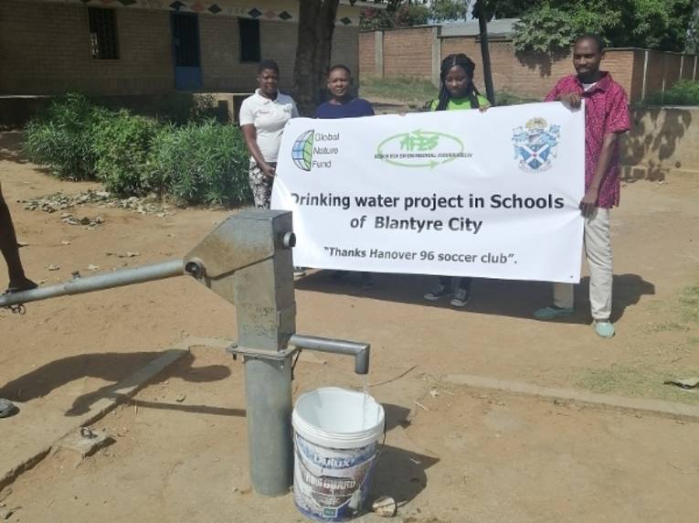 Menschen auf einem Schulgelände mit einem Plakat zum Brunnenprojekt. Im Vordergrund ist die reparierte Wasserpumpe.