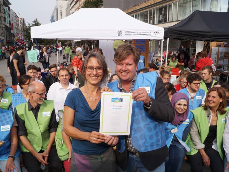 Susanne Wildermann vom Agenda21- und Nachhaltigkeitsbüro und Carsten Elkmann von TransFair e.V.  präsentieren vor Aktiven am Kröpke die Fair Trade Town Urkunde für Hannover