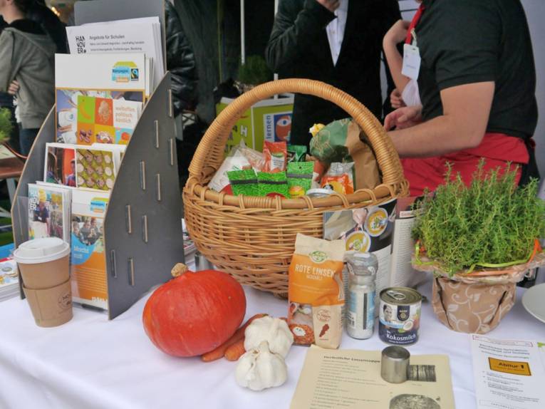 Auf einem Tisch sind Info-Broschüren in einem Ständer ausgestellt. Außerdem steht ein Korb mit fair gehandelten Lebensmitteln auf dem Tisch sowie weitere fair gehandelte Produkte wie Kürbis, Kokosmilch oder Linsen.