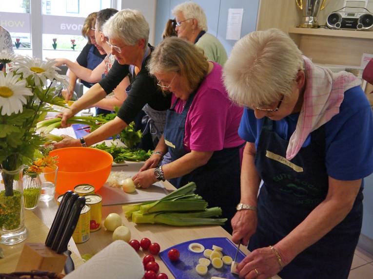 Frauen mit Küchenschürzen stehen an einer großen Arbeitsplatte und bereiten Gemüse zum Kochen vor.