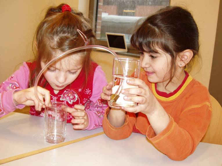 Zwei Mädchen experimentieren mit zwei Wassergläsern. Eines ist gefüllt, das andere ist leer, dazwischen ist ein Verbindungsschlauch.