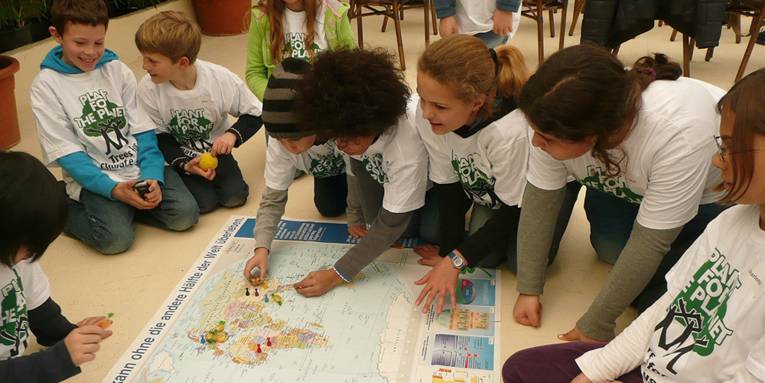 Kinder knien um eine große Weltkarte und markieren mit kleinen "Mensch-ärger-Dich-nicht"-Spielfiguren Waldgebiete