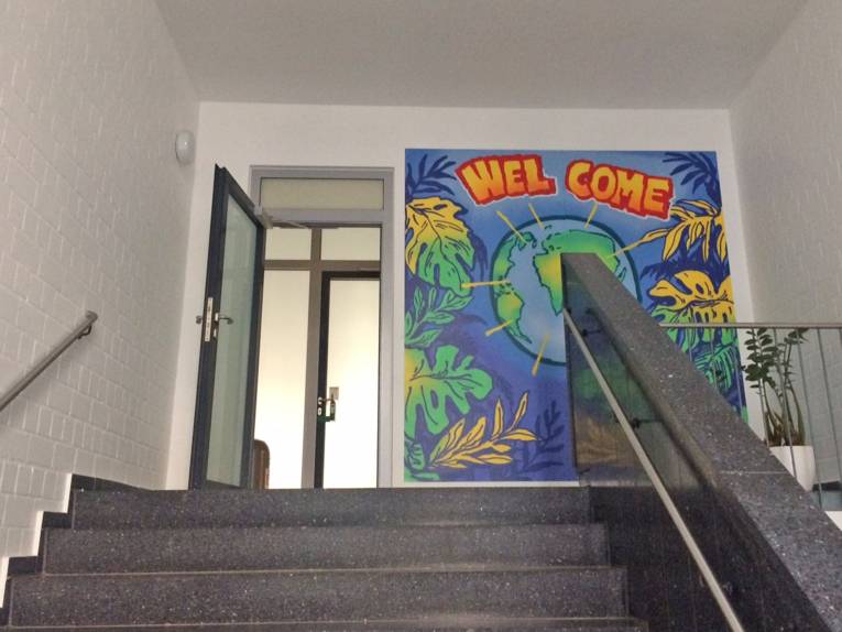 Eine Treppe führt zu einer offenen Tür, neben der eine bunte "Welcome"-Zeichnung hängt.