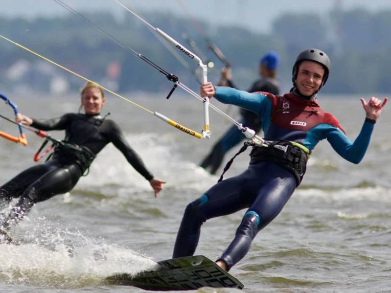 Ein junger Mann und eine junge Frau in Neoprenanzügen surfen auf ihren Boards und Kites über das Wasser.