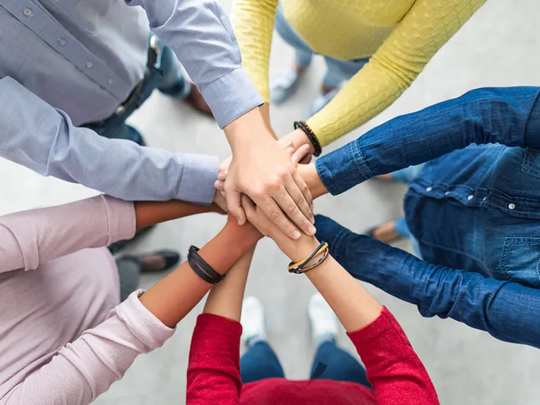 F&uuml;nf Menschen stehen im Kreis und haben die H&auml;nde &uuml;bereinander gelegt.