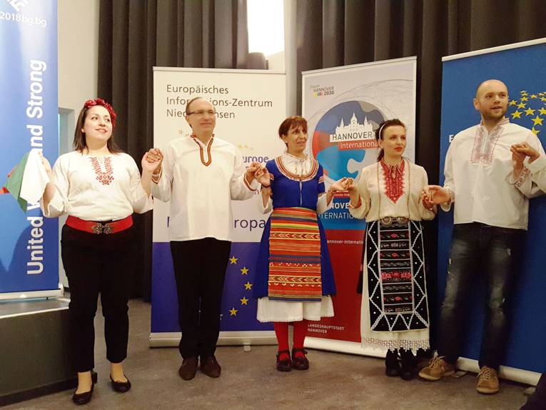 Drei Tänzerinnen und zwei Tänzer in bulgarischen Trachten haben sich an den Händen gefasst und tanzen auf einer Bühne.