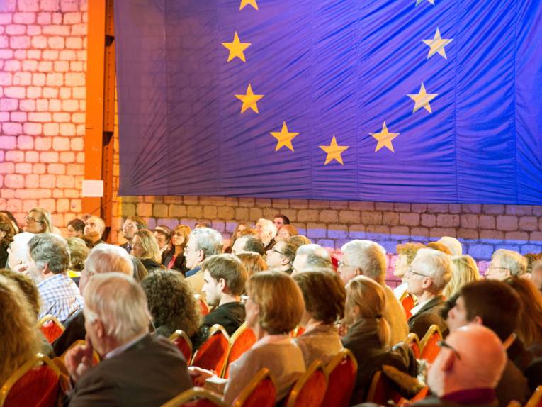 Vor dem Hintergrund einer Europa-Fahne sitzen zahlreiche Zuhörerinnen und Zuhörer in einem Saal.