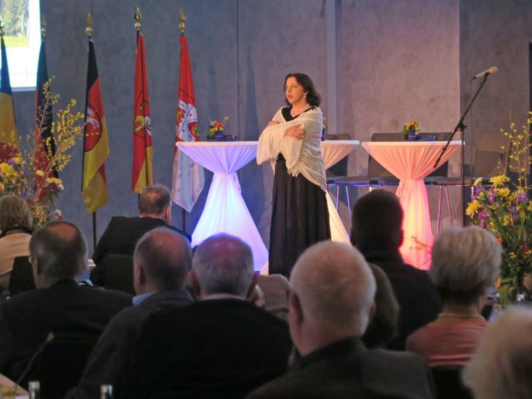 Eine Schauspielerin auf der Bühne rezitiert mit verschränkten Armen. Im Hintergrund fünf unterschiedliche Flaggen, im Vordergrund das Publikum, das sitzend dem Vortrag zuschaut.