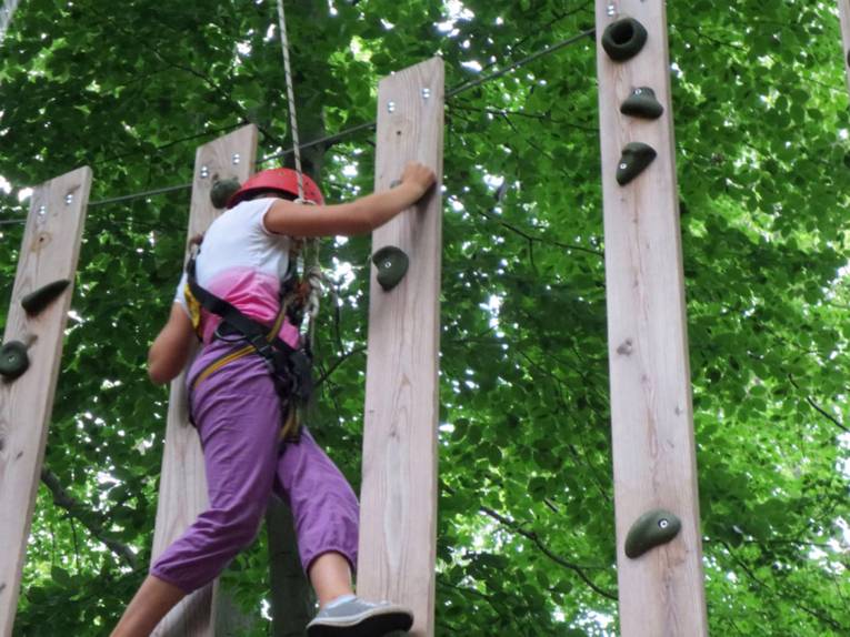 Ein Kind balanciert durch Seile und Helm abgesichert an einer Holzwand