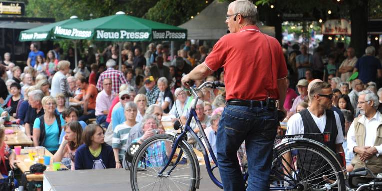 Auf der Bühne des Sommerfestes der Neuen Presse steht ein Auktionator und versteigert ein Fahrrad vor den auf Bänken sitzendem Publikum.