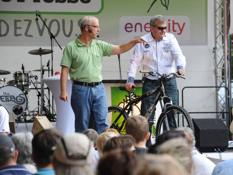 Auf der Bühne des Sommerfestes der Neuen Presse steht ein Auktionator und versteigert ein Fahrrad, das von einem Herrn gehalten wird, damit das Publikum den Versteigerungsgegenstand sehen kann.