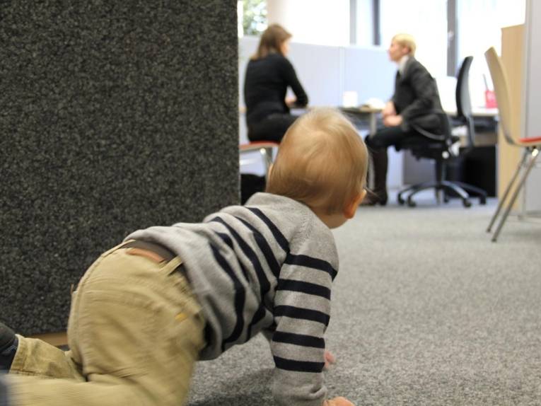 Kleines Kind krabbelt im Vordergrund, während sich die Mutter im FamilienServiceBüro beraten lässt