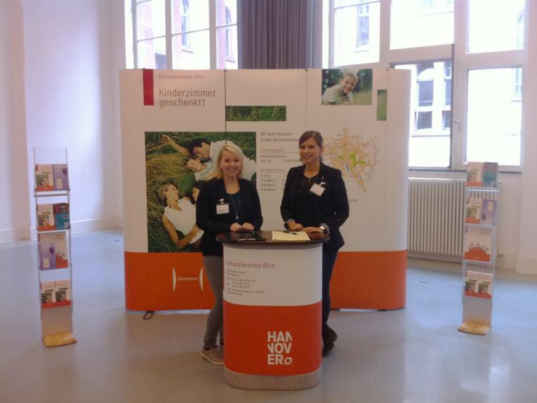 Zwei Mitarbeiterinnen am Informationsstand des Einfamilienhaus-Büros, dahinter eine Stellwand mit der Aufschrift "Kinderzimmer geschenkt!"