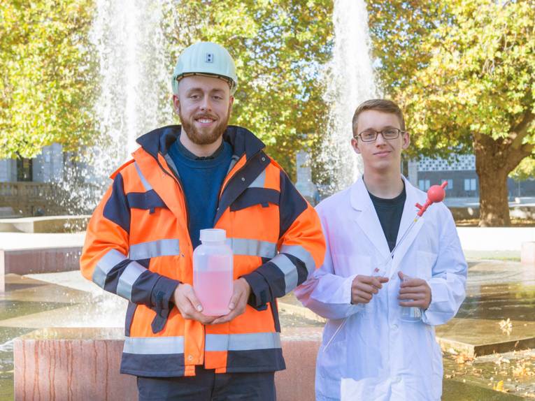 Zwei junge Männer in Berufskleidung, einmal mit Schutzhelm und Jacke und einmal mit Laborkittel, vor einem Springbrunnen 