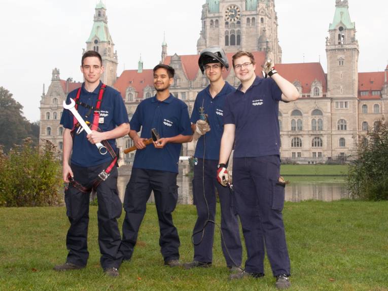 Vier Auszubildende in Arbeitskleidung und mit Arbeitsgerät vor dem Neuen Rathaus Hannover