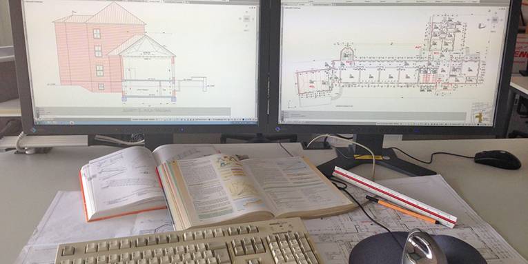 Zwei Monitore, die Anschnitt und Gebäudepläne zeigen, und ein Schreibtisch, auf dem ein Bauplan, eine Tastatur, Bücher, Stift und Lineal liegen