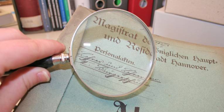 Eine Hand mit einer Lupe über einer historischen Personalakte, die in alter deutscher Schrift beschriftet ist