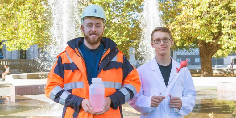 Zwei junge Männer in Berufskleidung, einmal mit Schutzhelm und Jacke und einmal mit Laborkittel, vor einem Springbrunnen 