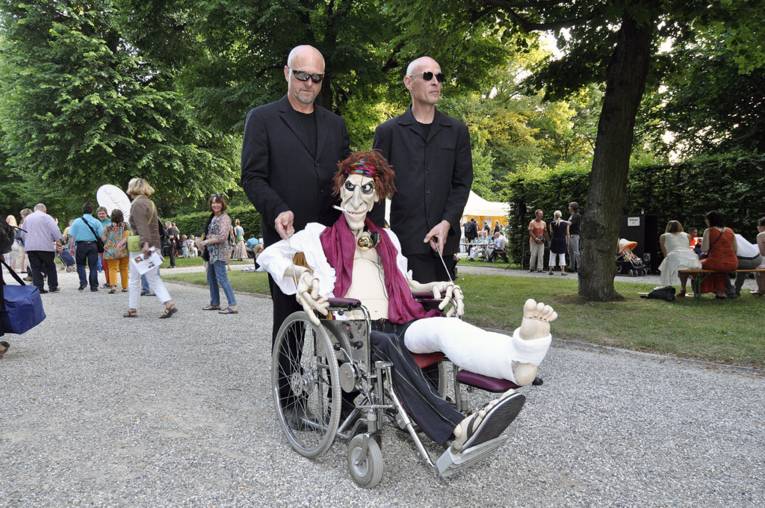 Zwei Männer schieben eine Puppenfigur im Rollstuhl durch den Park.