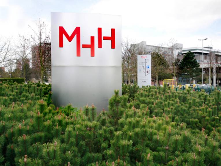 Stele mit dem Schriftzug MHH inmitten von Bäumen, daneben ein Parkplatz.