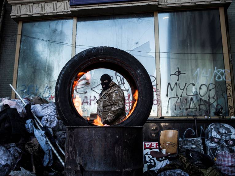 Mann mit Sturmhaube durch brennenden Autoreifen hindurch gesehen