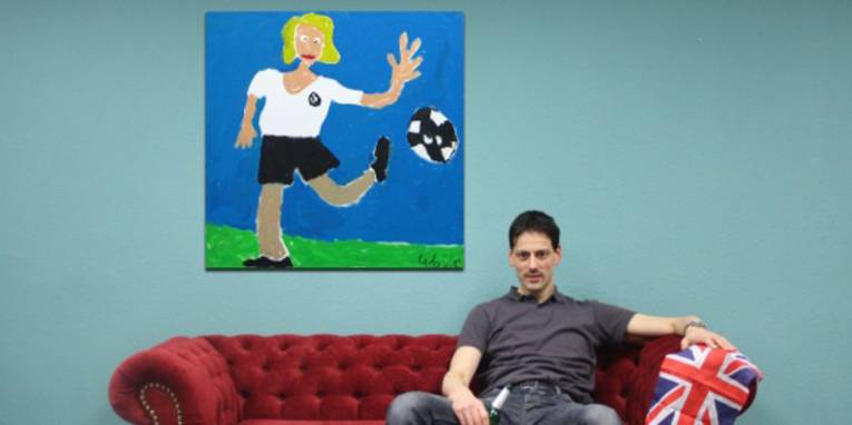 Mann sitzt auf einem Sofa mit einer Englandfahne in der Hand. Hinter ihm hängt ein Bild an der Wand, das eine Fußballerin in deutschem Trikot zeigt.