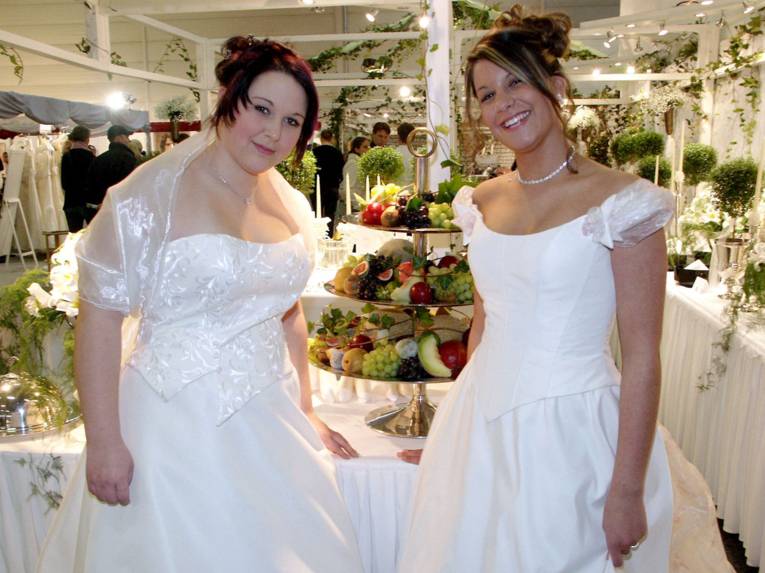 Zwei junge Frauen in Brautkleidern vor einem Etagere mit Obst 