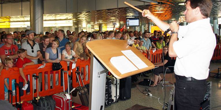 Mann mit Holzhammer steht an einem Pult. Davor sind Koffer und Reisetaschen sowie hinter einer Absperrung viele Zuschauer in einem Flughafenterminal.
