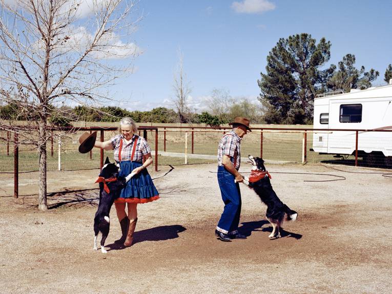 Ein älterer Mann und eine ältere Frau in Western-Kleidung tanzen mit ihren Hunden vor einem Wohnwagen.