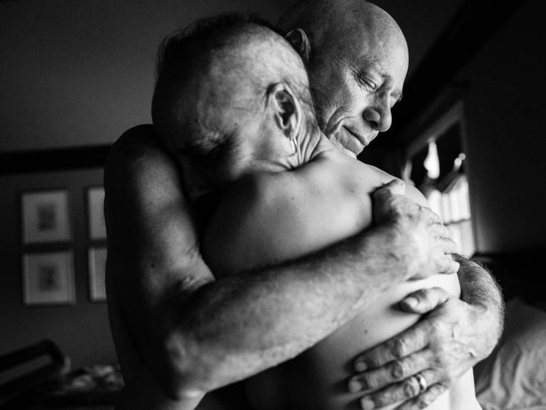 Zwei kahlköpfige Männer mit freiem Oberkörper in einem Krankenzimmer umarmen sich.