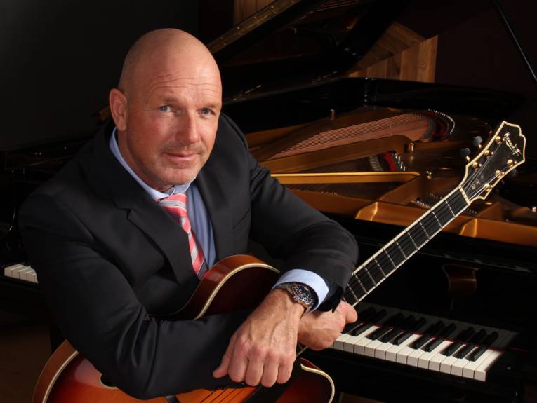 Ein Mann sitzt mit einer Gitarre auf dem Schoß vor einem Flügel.