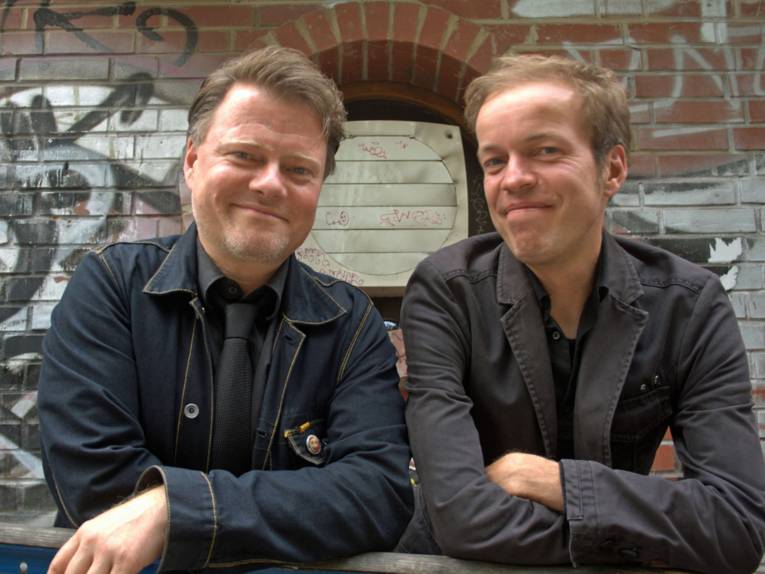 Zwei lachende Männer in dunklen Jacken vor einer Steinmauer.