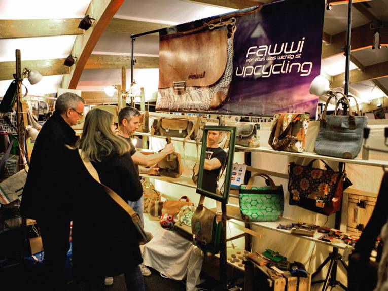 Zwei Männer und eine Frau an einem Stand mit Taschen.