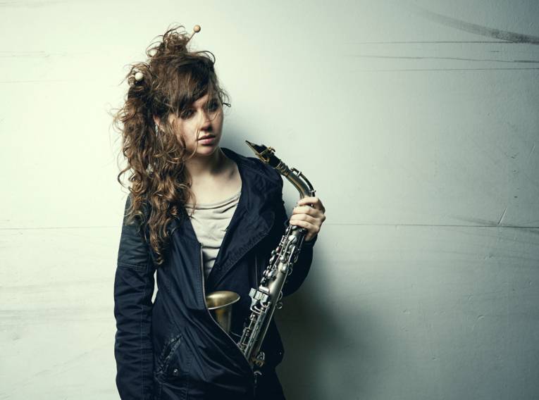 Eine junge Frau mit wilden langen Locken hält ein Saxophon in der Hand.