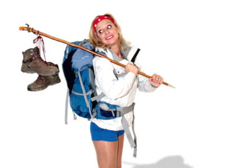 Eine Frau in kurzen Hosen mit Rucksack und einem Stab mit Wanderschuhen über der Schulter.