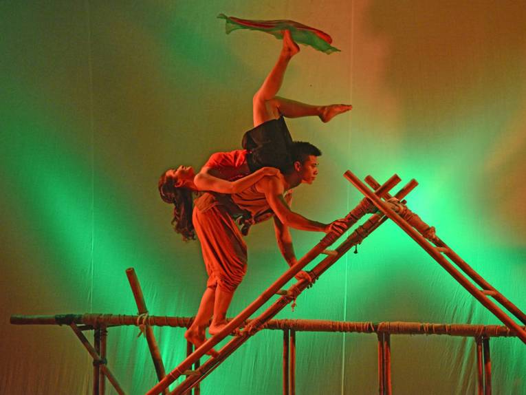 Mann trägt Frau auf dem Rücken, Frau balanciert ein Tuch auf dem Fuß.