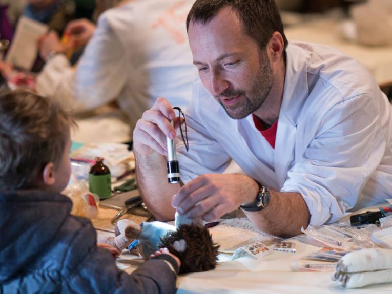 Mann in weißem Kittel untersucht ein Plüschtier unter Einsatz einer Taschenlampe