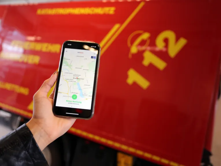 Jemand hält ein Handy, im Hintergrund ein Feuerwehrauto