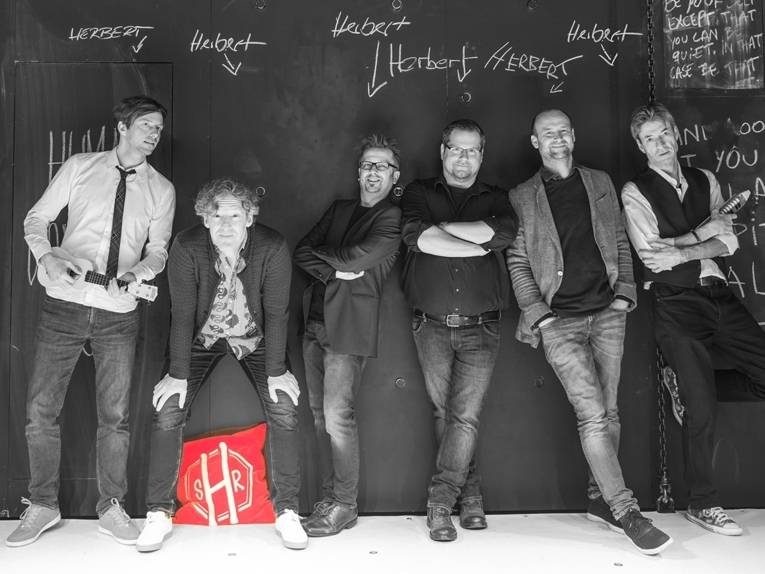 Sechs Männer stehen an einer Wand. Auf alle zeigt jeweils ein Pfeil mit der Beschriftung " Herbert".