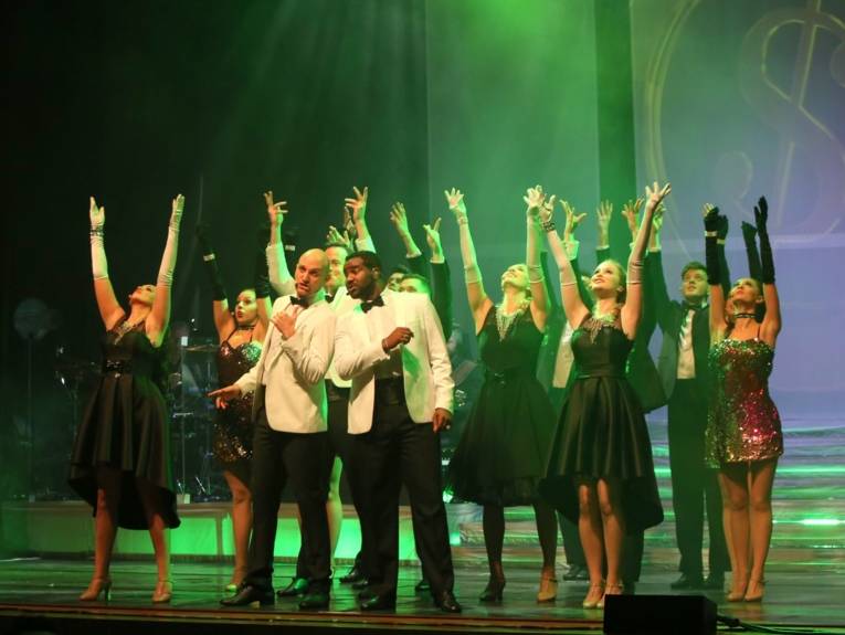 viele Tänzer und Tänzerinnen auf einer Bühne. Sie halten die Hände in die Luft.