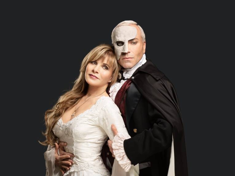 Frau mit blonden Harren und einem Kleid, wird von einem Mann mit Maske gehalten.