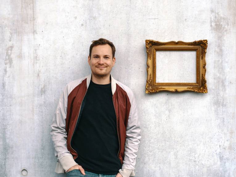 Ein Mann steht vor einer Wand. An der Wand hängt ein goldener Bilderrahmen.