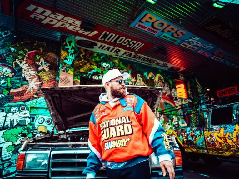 Ein Mann mit Bart und einem bunten Pullover steht vor einer Graffiti Wand. Ein Auto steht hinter ihm und er lacht zur Seite.