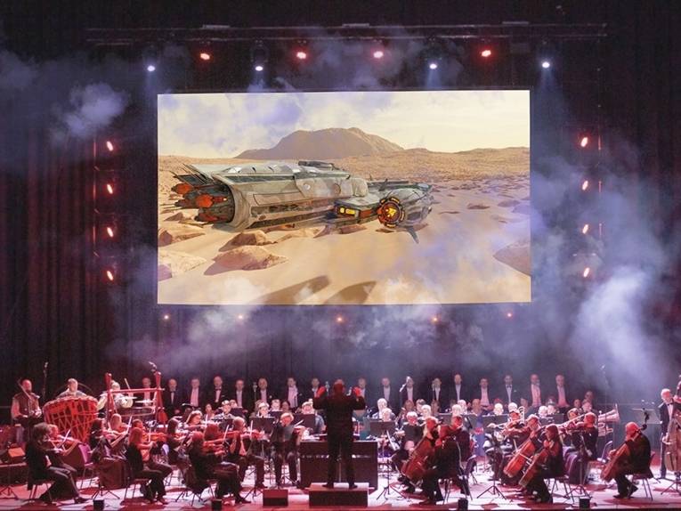 Großes Orchester auf einer Bühne, im Hintergrund Bild einer Wüstenlandschaft mit einem Raumschiff