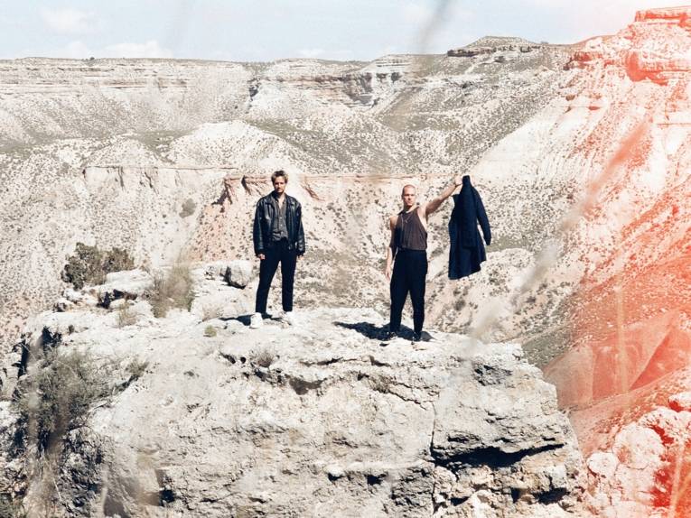 Zwei Männer stehen auf einem Felsbrocken mitten in der Natur, umrahmt von Felsen. Der eine Mann hält seinen Mantel in die Luft.