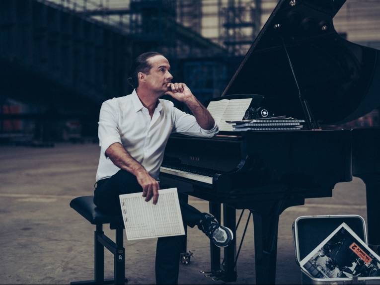 Ein Mann sitzt vor einem Flügel und denkt nach, mit Notenblättern in der Hand.