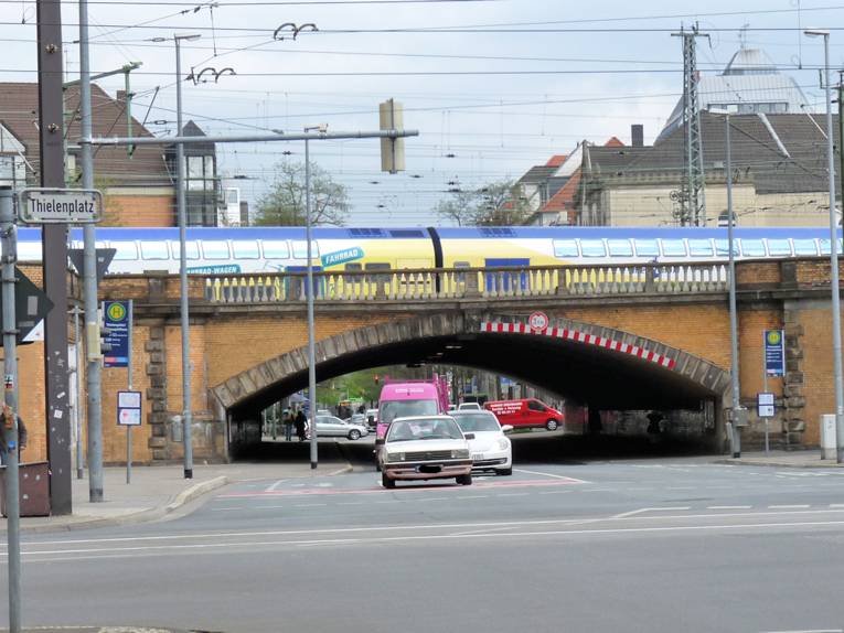 Eisenbahnbrücke innerorts mit Zug und Kraftfahrzeugen.