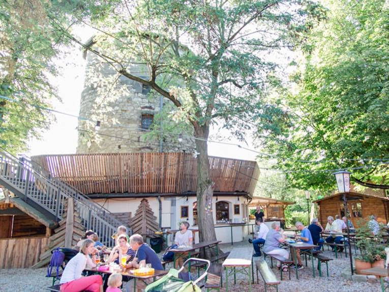 Menschen sitzen auf Bänken an Tischen vor einen historischen Turm.
